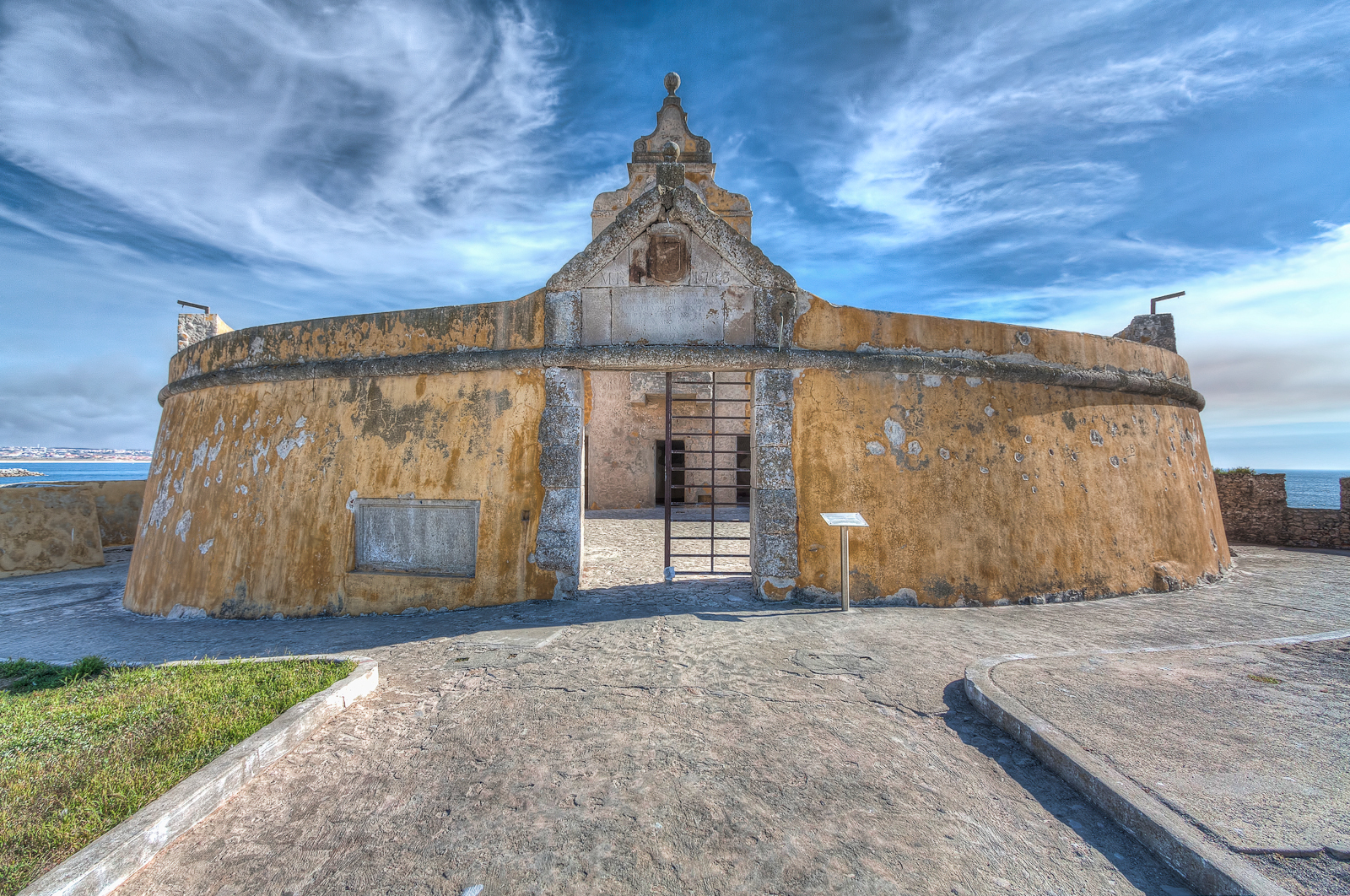 Entrada principal do Fortim Redondo, Fortaleza de Peniche, com a sua pedra e tinta amarela descascada, contra um céu azul riscado por nuvens brancas.