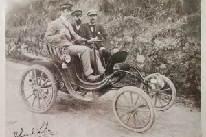 Fotografia a preto e branco, em tons de sépia, da primeira subida à Serra da Estrela em automóvel, protagonizada em 1906 por João Alves da Silva, personalidade marcante da história da Covilhã, assinada pelo próprio, fotografado ao volante, e com a inscrição "1º Automóvel que entrou na Covilhã em 1901". Estão presentes mais dois indivíduos, apeados, não identificados.