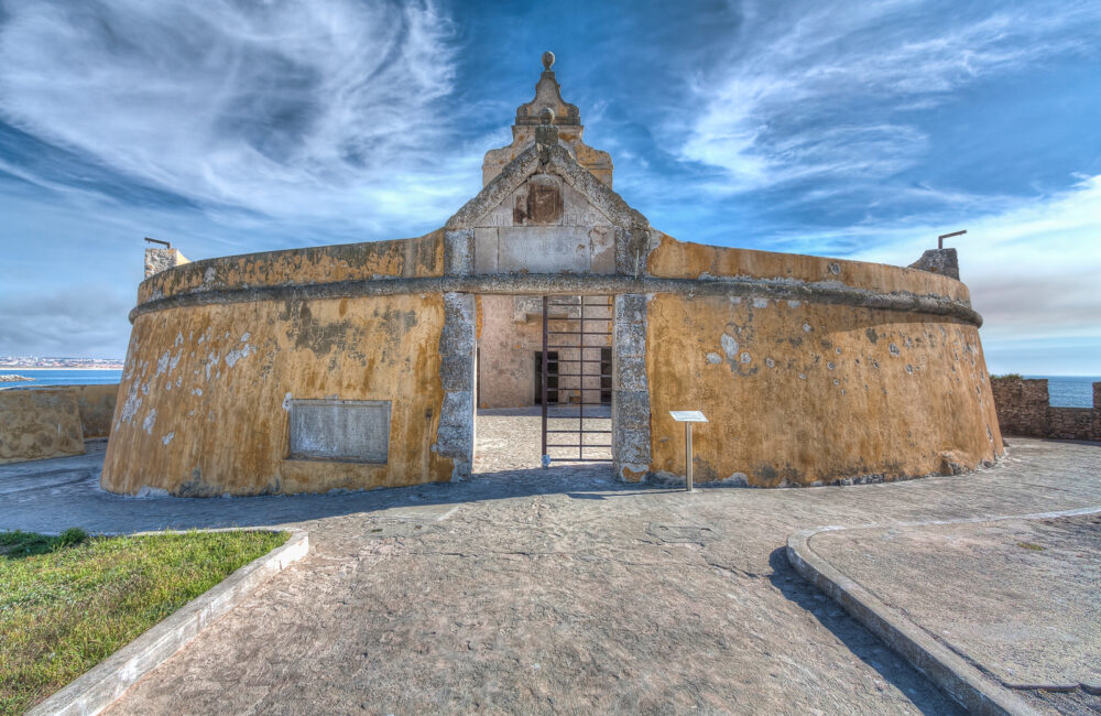 Entrada principal do Fortim Redondo, Fortaleza de Peniche, com a sua pedra e tinta amarela descascada, contra um céu azul riscado por nuvens brancas.