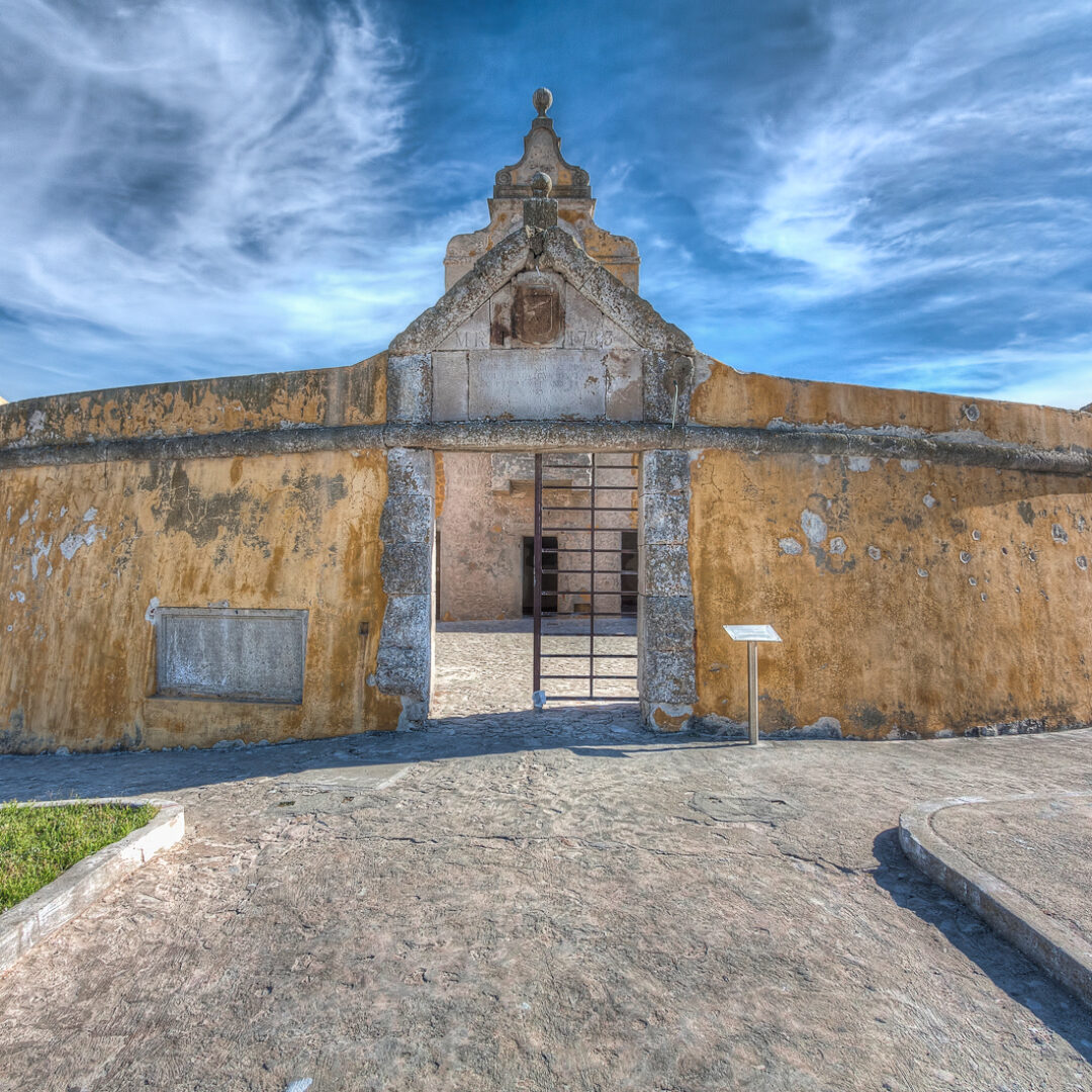 Entrada principal do Fortim Redondo, Fortaleza de Peniche, com a sua pedra e tinta amarela descascada, contra um céu azul riscado por nuvens brancas.